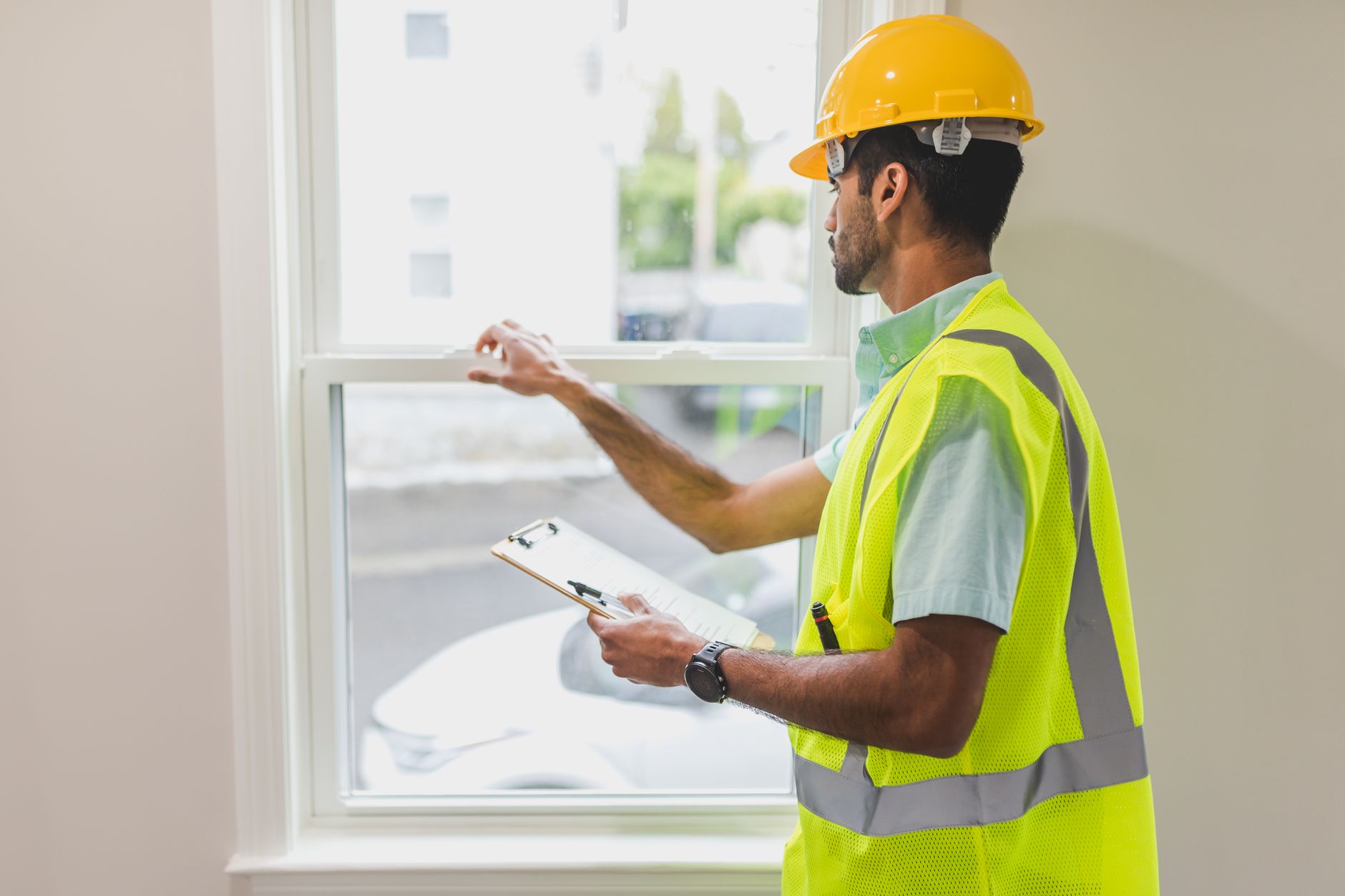 A construction worker in a safety vest and hard hat inspects a window indoors, holding a clipboard in Austin, TX